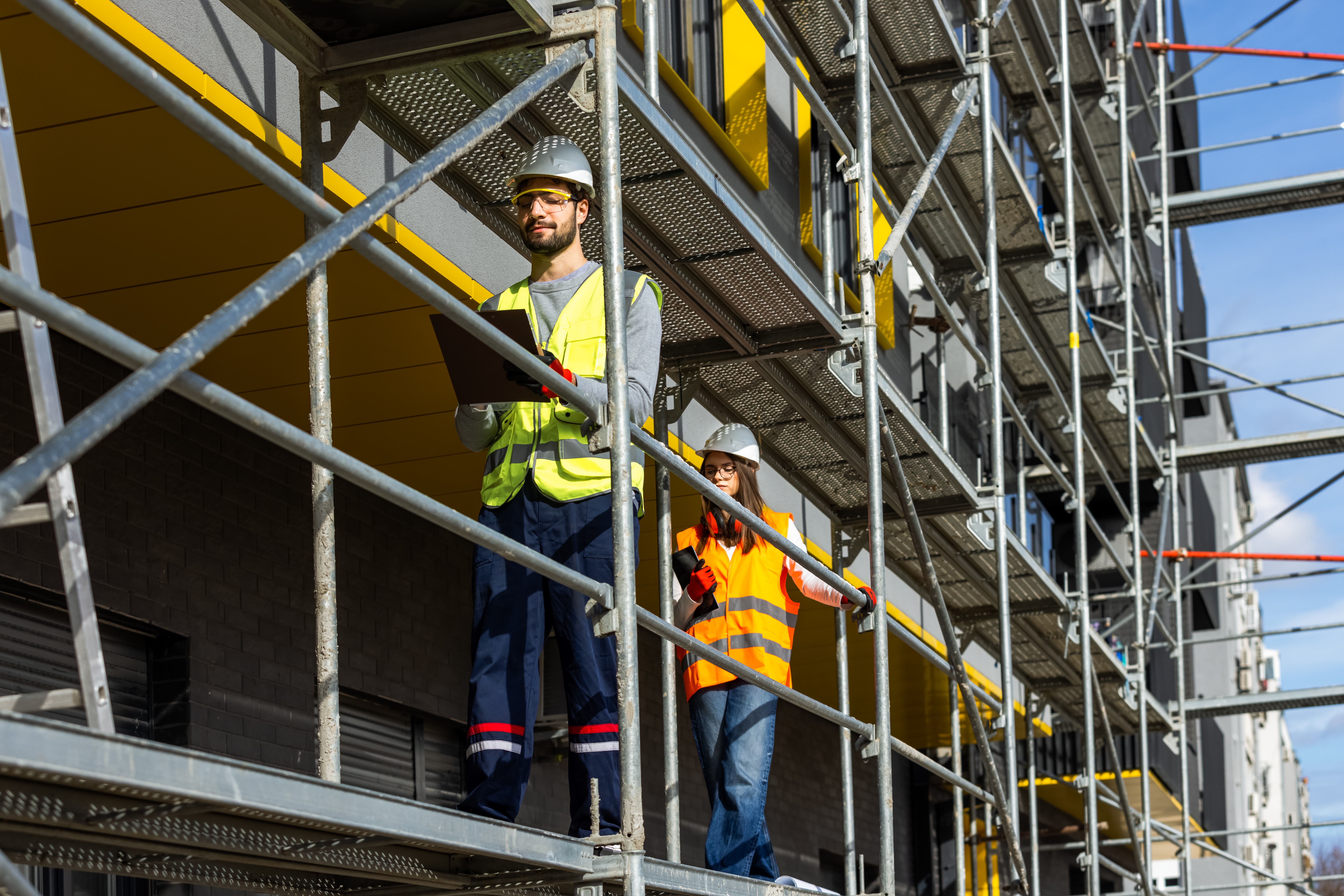 Two professional engineers in safety gear walking along construction scaffolding while surveying the building project. This image effectively captures the dynamic nature of field inspections and modern construction management.